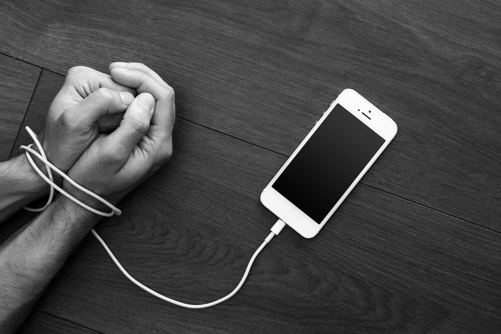cellphone on a table with the charging cord wrapped around a person's wrists as a concept image for cellphone addiction