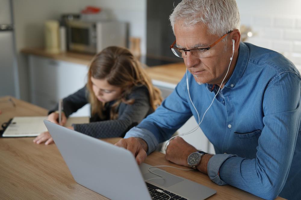 remote worker listening in on a meeting in his kitchen with his daughter working on homework by his side